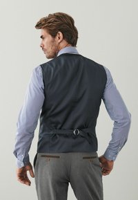 Dunkelblaues Weste mit glatter Textur, verstellbarem Rückengurt, über einem gestreiften Hemd getragen. Graue Hose mit braunen Knopfakzenten.