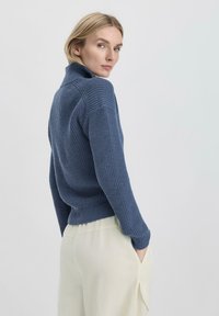 Blauwe gebreide trui met hoge kraag en ribgebreide mouwen, gedragen over crèmekleurige wijde broekspijpen, met een textuurdesign van de stof.