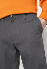 Personne portant un pantalon gris foncé avec un bouton et une fermeture éclair, et un haut orange à manches longues avec une main dans la poche.