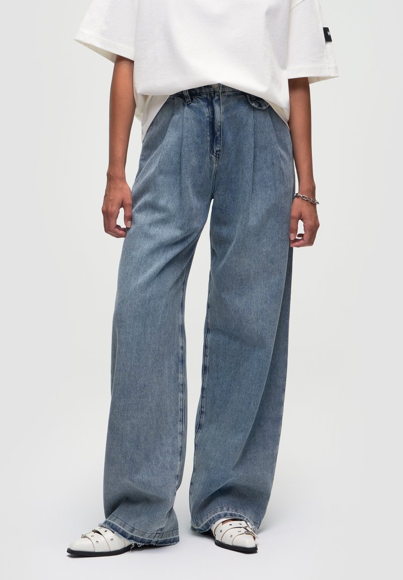 Jeans de mezclilla azul claro, de tiro alto y pernera ancha, con plisados en la parte delantera y bordes rematados, combinados con zapatos blancos sin cordones.