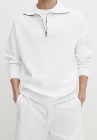 Persona vestita con un pullover bianco a mezza zip e pantaloni bianchi, con le mani nelle tasche, contro uno sfondo neutro.