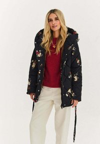 Čierna prešívaná vatovaná bunda s kvetinovou výšivkou, kapucňou a zapínaním na cvoky, nosená cez červený sveter a svetlé nohavice.