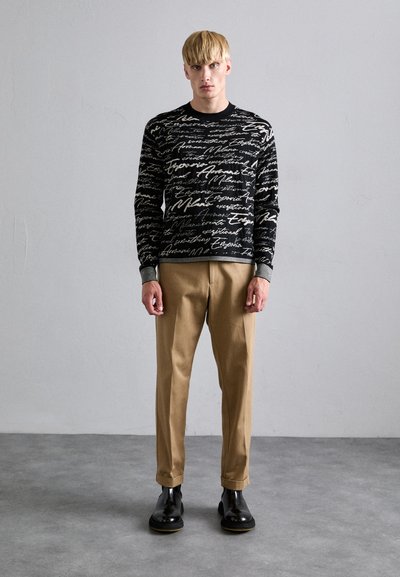 Sudadera negra con texto en cursiva blanca, de mangas largas y puños acanalados; combinada con pantalones beige y botas negras. Textura suave y diseño ajustado.
