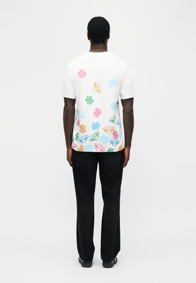 Άτομο με την πλάτη, φοράει λευκό T-shirt με πολύχρωμα σχέδια δολαρίων και διαμαντιών, συνδυασμένο με μαύρο παντελόνι και μαύρα παπούτσια.