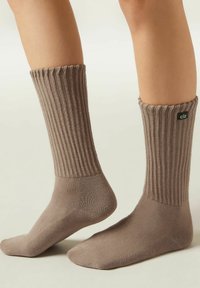 Braune, gerippte Socken mit einem strukturierten Bündchen, hergestellt aus weichem Stoff. Enthält ein schwarzes Logoetikett auf der Oberseite und einen glatten Fußbereich.