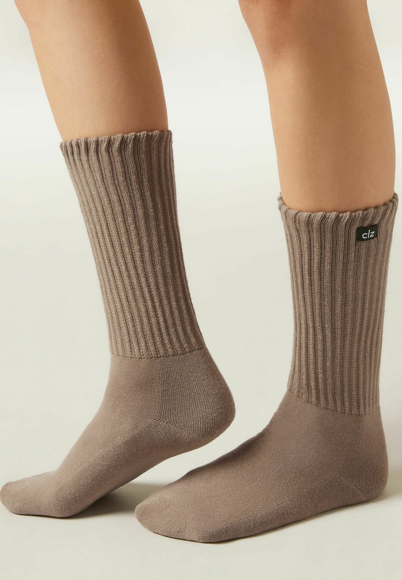 Braune, gerippte Socken mit einem strukturierten Bündchen, hergestellt aus weichem Stoff. Enthält ein schwarzes Logoetikett auf der Oberseite und einen glatten Fußbereich.