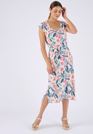 Mujer con vestido midi sin mangas con estampado de hojas tropicales de colores y sandalias doradas, de pie con una mano tocándose el cabello.