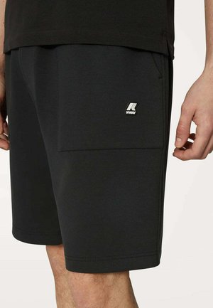 Shorts neri alla lunghezza del ginocchio con una tasca che presenta un piccolo logo K-Way bianco sulla coscia destra, indossati con una maglietta nera.