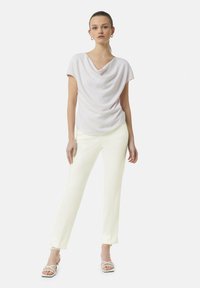 Korte mouwen, gedrapeerde top in lichtroze stof, gecombineerd met op maat gemaakte, recht gesneden broeken in het wit. Draagt witte sandalen met banden.