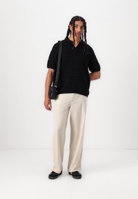 Polo shirt negru tricotat cu mâneci scurte, asortat cu pantaloni largi de culoare bej deschis și încălțăminte neagră. Include o geantă mică neagră de umăr.