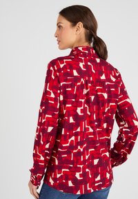 Camicia a maniche lunghe rossa e bianca con motivo. Presenta un colletto e un dettaglio di piega sul retro. Tessuto morbido con un design astratto.