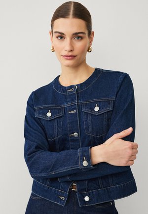 Woman with slicked-back hair wearing gold hoop earrings and a dark blue denim jacket with metal buttons, standing with arms crossed.
