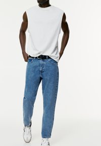 White sleeveless t-shirt, blue denim jeans with a relaxed fit, accented by a black belt and white sneakers with black details.