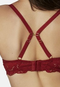 Soutien-gorge en dentelle rouge avec des bretelles croisées réglables aux bords festonnés, des éléments en or et des motifs floraux texturés.