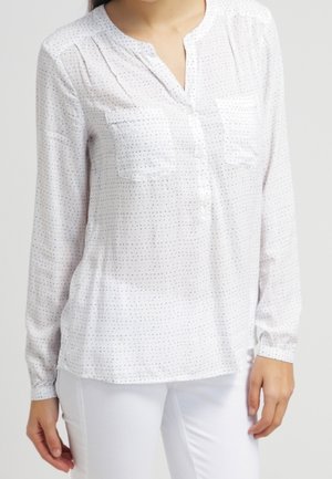 Vrouw draagt een wit blouse met lange mouwen, voorzien van kleine zwarte stippen en twee borstzakken, gecombineerd met een witte broek, tegen een effen achtergrond.