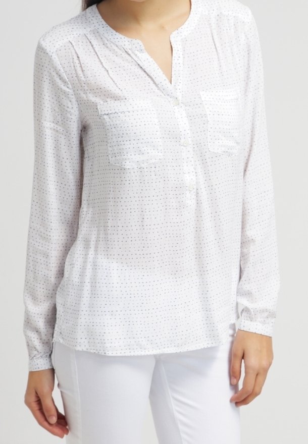Femme portant un chemisier blanc à manches longues avec de petits pois noirs et deux poches poitrine, associé à un pantalon blanc, sur un fond uni.