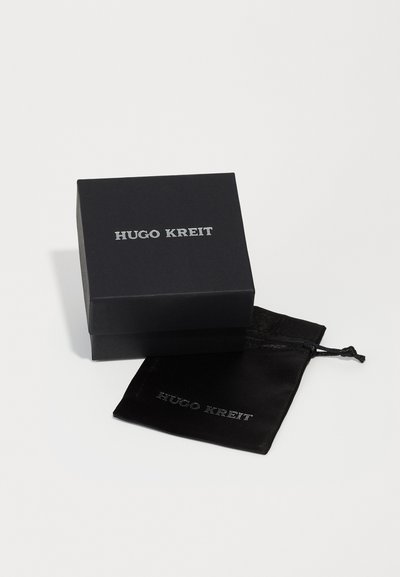 Boîte à bijoux noire avec le texte argenté "HUGO KREIT" sur le dessus, accompagnée d'un pochon en satin noir portant le même texte, le tout élégant et rectangulaire.