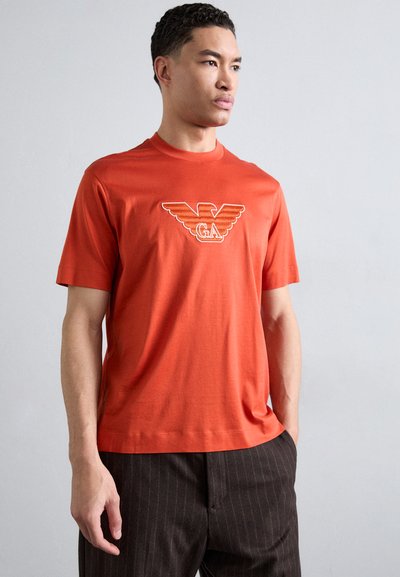 Oranžinė marškinė su apvalia apykakle, pagaminta iš minkštos medžiagos; turi kontrastingą baltą grafiką ant krūtinės.