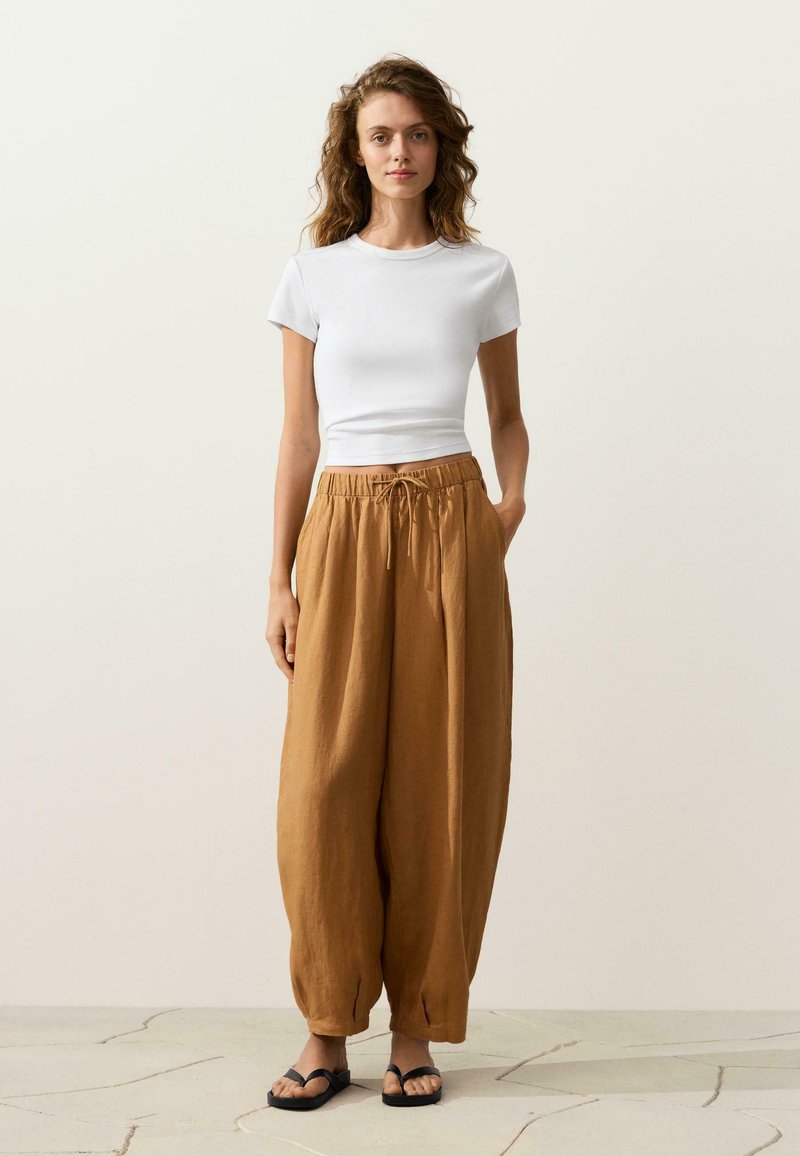 Femme portant un haut blanc ajusté à manches courtes, un pantalon marron ample avec cordon de serrage et des sandales noires, debout sur un sol carrelé clair.
