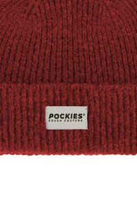 Piros, kötött sapka puha, textúrált anyagból. Bordás mintázattal rendelkezik, és egy fehér címkén fekete felirattal olvasható: "POCKIES COUCH COUTURE."