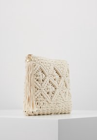 Kremowa torba makramowa z misternymi wzorami w diamenty, charakteryzująca się teksturowaną powierzchnią i dekoracyjnym chwostem po boku. Złote detale na suwak.
