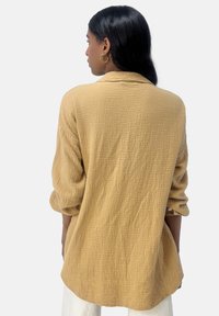 Langarmshirt aus strukturiertem Senfstoff, mit lockerer Passform und einer Knopfdetails am Ärmelbund. Die Rückansicht zeigt das drapierte Design.