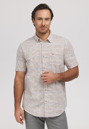 Mężczyzna ubrany w beżową, krótko rękawową koszulę w kratę z guzikami, stojący na tle jednolitego jasnego tła, patrzący w kamerę.