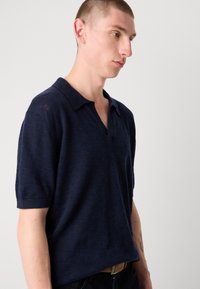Mørkeblå polo t-skjorte med en kort, litt oversized passform, har en klassisk krage og et teksturert strikket mønster.