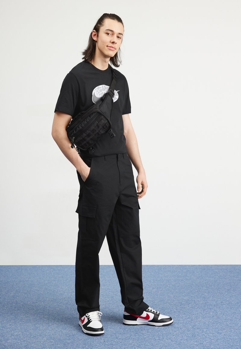 T-shirt noir avec un motif graphique, pantalon cargo noir et baskets Nike noires/rouges. Comprend un sac bandoulière noir avec un design texturé.