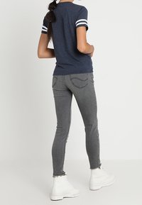 Kvinna som står med ryggen mot oss, klädd i t-shirt med marinblå ränder, urtvättade grå skinny jeans med fransade nederkanter och vita snörstövlar.