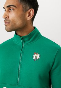 Sweatshirt zippé vert avec col montant, arborant le logo brodé des Boston Celtics sur la poitrine, fabriqué en matériau doux et extensible.