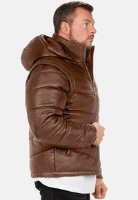 Giubbotto imbottito in pelle marrone con un cappuccio spesso, chiusura con zip e texture trapuntata. Presenta tasche laterali e polsini a coste per il calore.