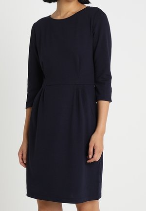 Frau trägt ein knielanges, marineblaues Kleid mit dreiviertel Ärmeln und tailliertem Schnitt vor einem schlichten Hintergrund.