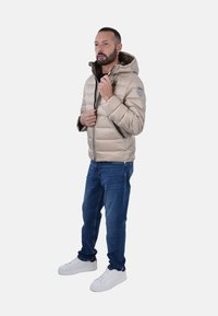 Giacca puffer beige con cappuccio, chiusura a zip, superficie testurizzata e una toppa sulla spalla. Abbinata a jeans blu e sneakers bianche.
