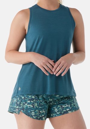 Vrouw met een mouwloos teal top en blauw-groene sportshorts met patroon, handen losjes samengevouwen voor zich.