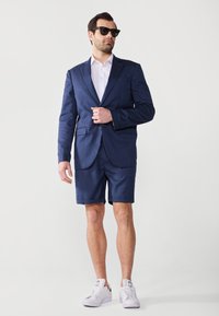 Veste de costume marine sur une chemise blanche, assortie à un short ajusté et des baskets blanches. Des lunettes de soleil noires complètent le look.