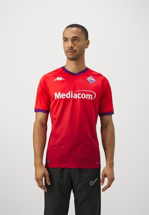 Uomo con maglia da calcio rossa della Fiorentina con logo Mediacom e pantaloni neri Nike, in piedi davanti a uno sfondo semplice.