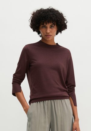 Vrouw met krullend haar, gekleed in een donkerbruine top met lange mouwen en lichtgrijze losse broek, handen in de zakken, tegen een effen achtergrond.