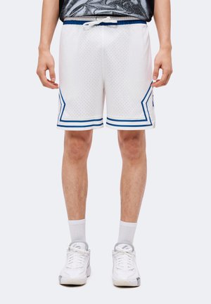 Person iført hvide basketballshorts med blå kant, hvide sokker og hvide sneakers, stående mod en ensfarvet baggrund.