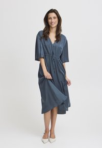 Blaues Satin-Kleid mit tiefem V-Ausschnitt, kurzen Ärmeln und gebundenem Taillengürtel. Der Rock ist fließend und hat einen unregelmäßigen Saum. Flache weiße Schuhe werden getragen.