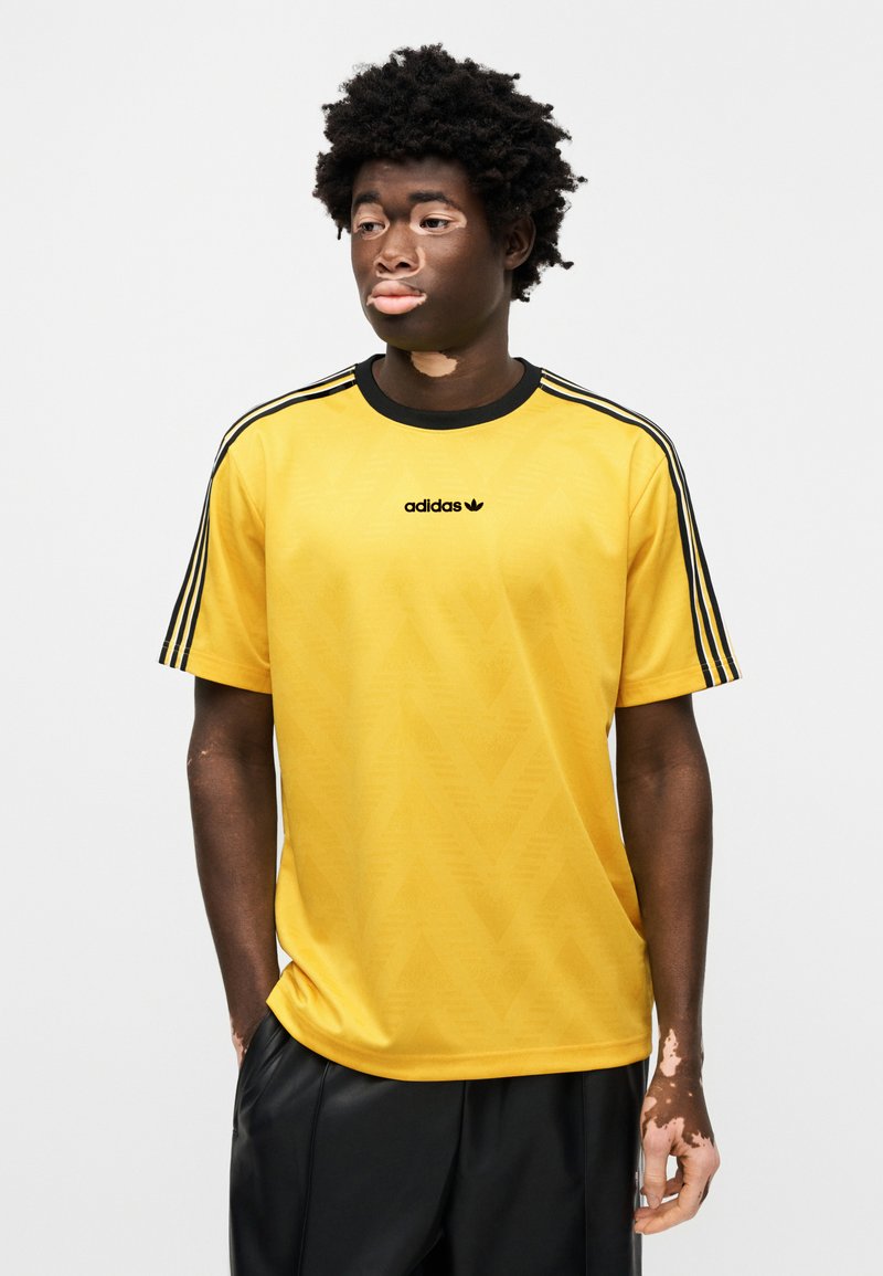 T-shirt jaune Adidas avec accents noirs et trois bandes le long des manches. Présente un motif texturé et un petit logo noir sur la poitrine.
