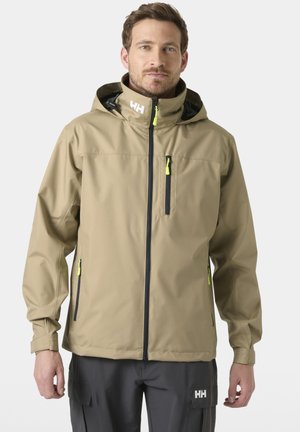Hombre con chaqueta impermeable color marrón claro con capucha y pantalones negros con logotipos de marca visibles, de pie frente a un fondo liso.