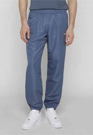 Mann iført blå treningsbukser med elastisk midje og ankler, kombinert med hvite joggesko, står foran en enkel lys bakgrunn.