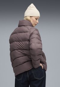 Kvinde iført en mauve puffjakke, mørke jeans og en cremet strikket hue, poserer med den ene hånd i lommen mod en grå gradient baggrund.