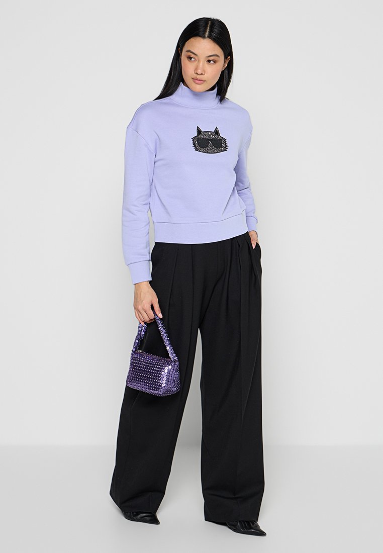 Sudadera lila con gráfico de gato, pantalones negros y un pequeño bolso de lentejuelas morado. Diseño de corte relajado y cuello alto. Zapatos negros.