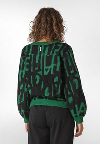 Svart och grön mönstrad tröja med lös passform, ribbade ärmslut och nederkant, med en etikett nära halsområdet. Mjuk textur och stickad material.