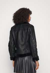 Zwarte leren jas met een bontkraag en manchetten; heeft een getailleerd ontwerp, gestikte panelen en een korte lengte. Plissé rok zichtbaar eronder.