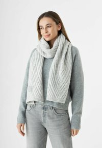 Vrouw die een lichtgrijze geribde gebreide sjaal draagt over een grijze trui en licht gewassen jeans, iets naar links kijkend met een zachte glimlach.
