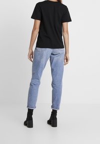 Sort kortærmet t-shirt, afslappet pasform, parret med lyseblå tætsiddende jeans. Modellen har sorte hælede ankelstøvler på. Simpelt baggrund.