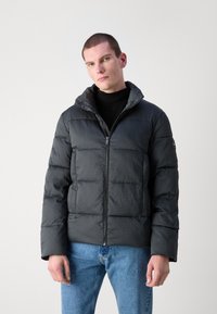 Giacca nera imbottita con zip frontale e cappuccio, caratterizzata da un motivo trapuntato e una texture liscia. Indossata sopra un dolcevita nero.